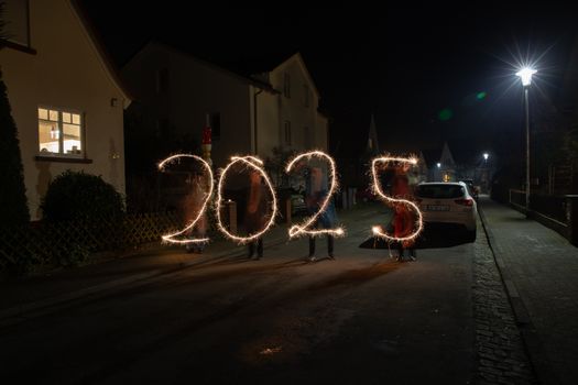 Sparkler light painting forming '2025' in a festive night street scene.